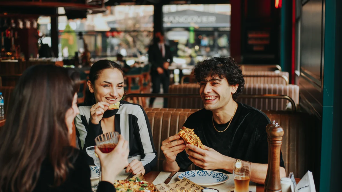 Group of friends enjoying food and drinks at a local Utica restaurant, including a man laughing with a sandwich, a smiling woman holding bread, and another woman holding a drink.
